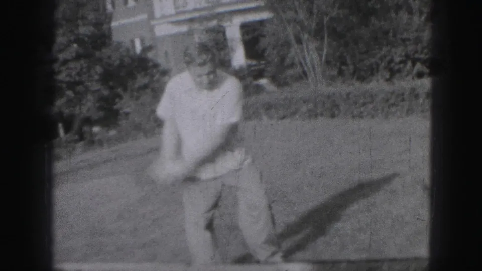 In 1975 Harrisburg home-movie footage, a man swings a golf club across a grassy yard with his shadow stretching to the side.