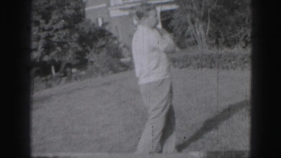 The man completes his golf swing in a grassy Harrisburg yard, with trees, a house exterior, and strong daylight shadow visible.