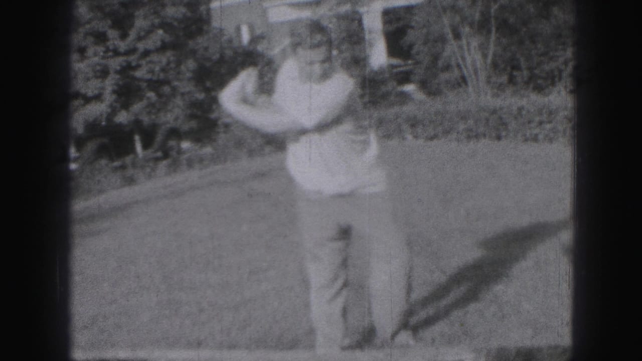 Man holding a golf club on a grassy lawn with trees and a house behind him.