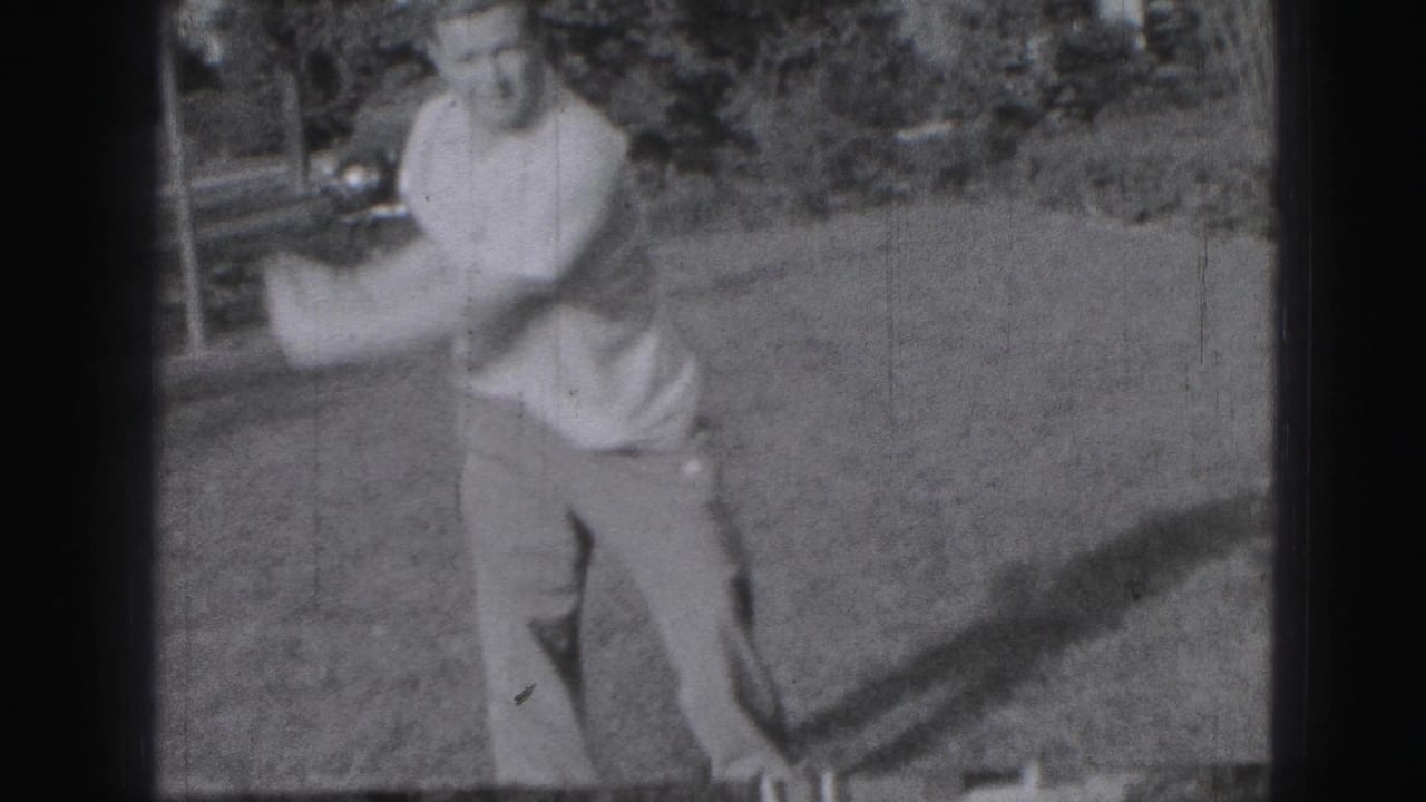 Adult man swinging a golf club on a grassy lawn with a parked car in the background.