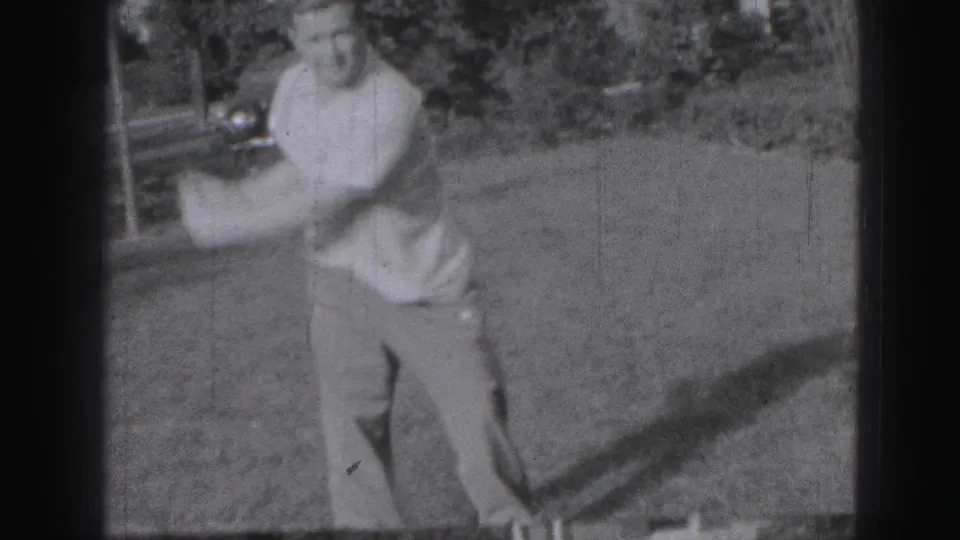 In the supplied 1975 Harrisburg home-movie context, an adult man begins a golf swing on a lawn with trees and a parked car behind him.