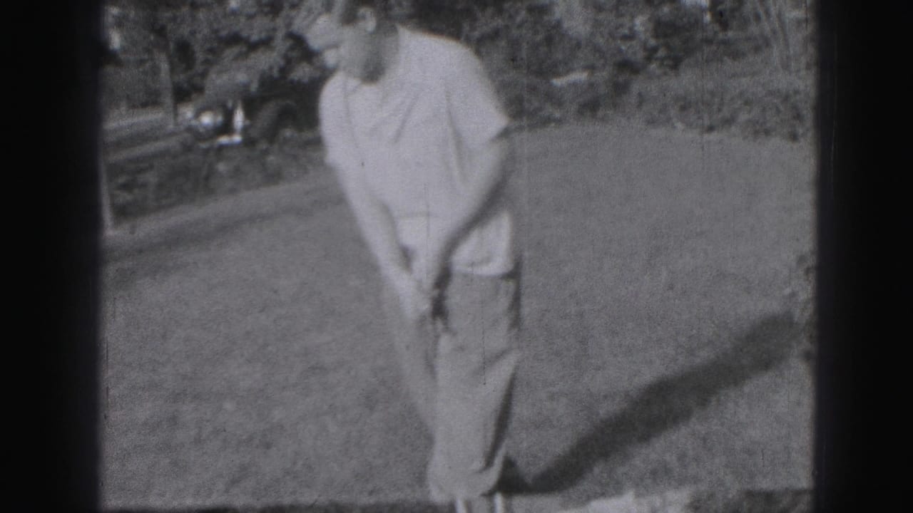 An adult golfer in light clothing stands over his club on a lawn in the 1975 Harrisburg home-movie footage, with a car and foliage behind him.