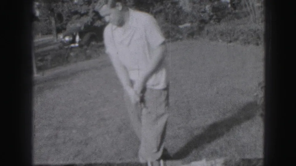 An adult golfer in light clothing stands over his club on a lawn in the 1975 Harrisburg home-movie footage, with a car and foliage behind him.