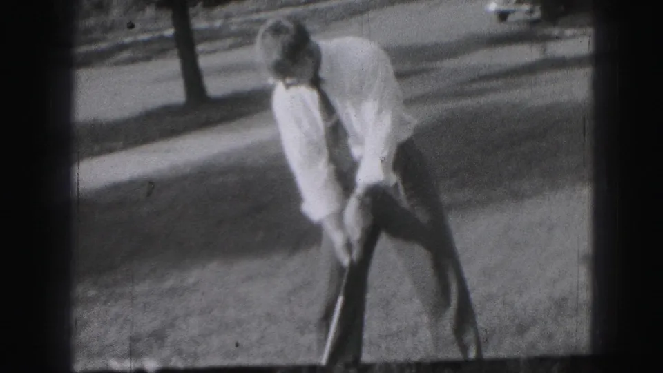 A 1975 Harrisburg home-movie frame shows a man in slacks, shirt, and tie leaning into a golf stance on grass.