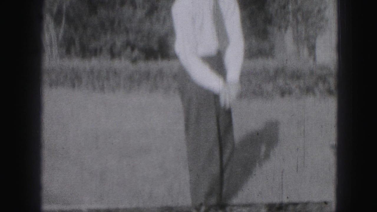 In 1975 Harrisburg home-movie footage, a young man stands on grass holding a golf club, partly cropped by the frame.