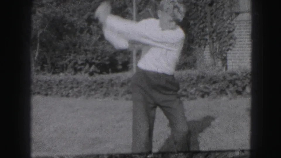A young man swings a golf club in a Harrisburg yard, with shrubs and a brick edge behind him in grainy 8mm footage.