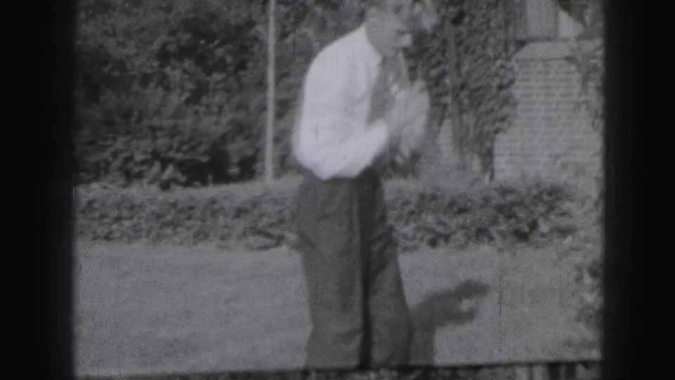 The man pauses or resets with the golf club near his shoulder, framed by lawn, shrubs, and a masonry background.
