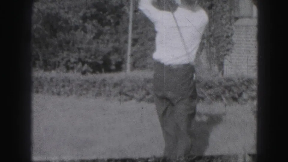 In 1975 Harrisburg home-movie footage, a man in a white shirt and tie stands on grass while starting a golf swing.