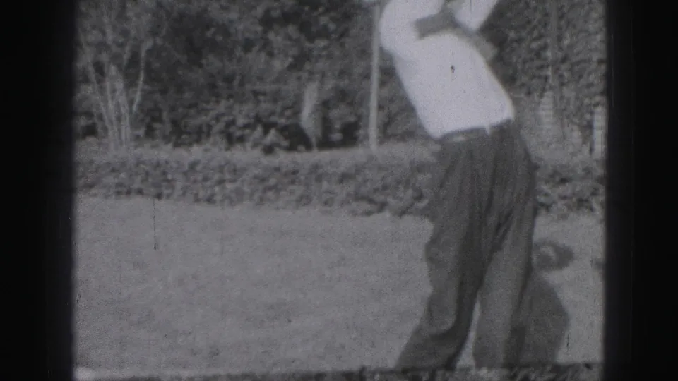 The man follows through on a golf swing, filmed in grainy 8mm from a residential yard setting in 1975 Harrisburg.