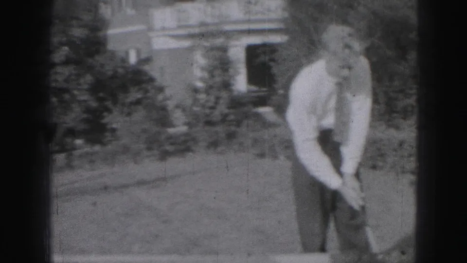 In Harrisburg, Pennsylvania home-movie footage from 1975, a man prepares a golf swing on a lawn near shrubs and a house exterior.