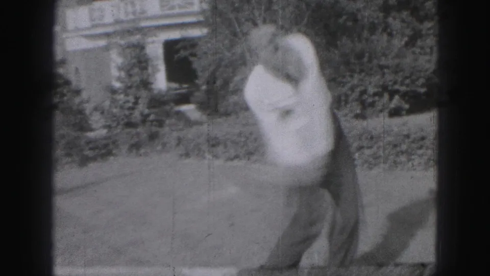The 1975 Harrisburg home-movie frame shows the man mid-swing, blurred by movement, with a hedge and house facade behind him.