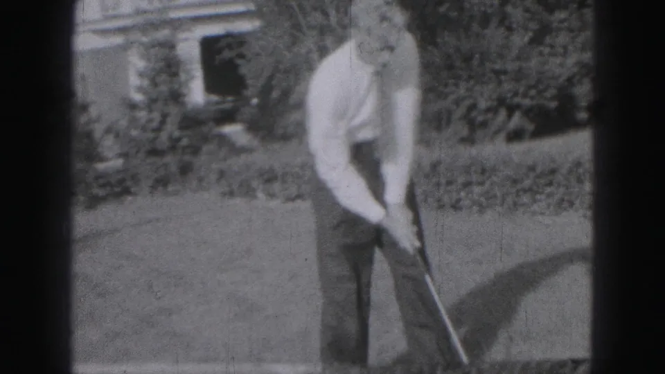 A 1975 Harrisburg backyard frame shows the man back in a golf stance, filmed in soft black-and-white 8mm texture.
