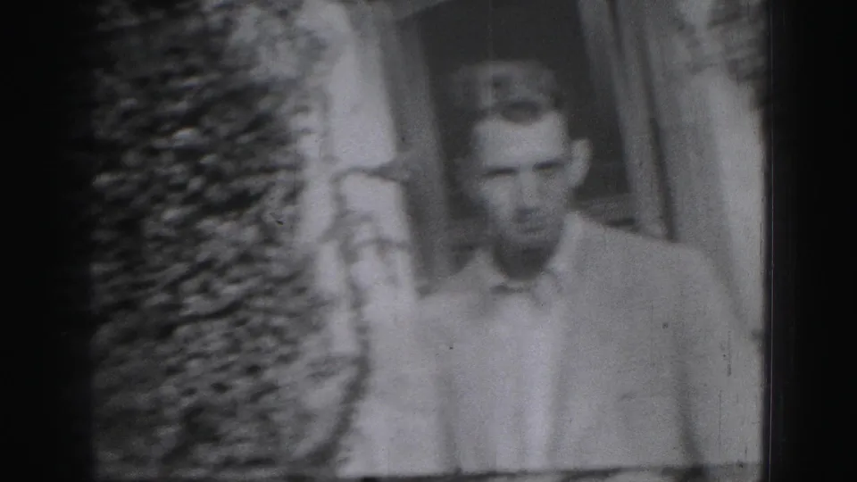 A young man in a light collared shirt appears outdoors near shrubs and a building in Harrisburg, Pennsylvania footage from 1975.
