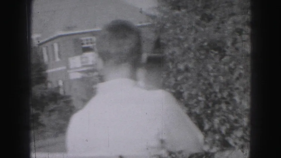 The back of a man in a light shirt is seen walking outdoors near shrubs and residential-looking buildings in 1975 home-movie footage.