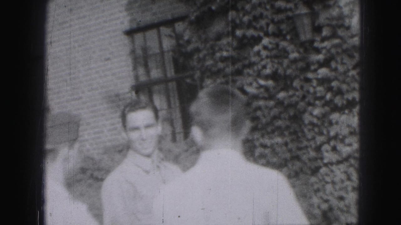 1975 Harrisburg home-movie frame showing a young man in a light shirt near another person beside a brick building and shrubs.