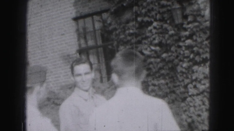 1975 Harrisburg home-movie frame showing a young man in a light shirt near another person beside a brick building and shrubs.
