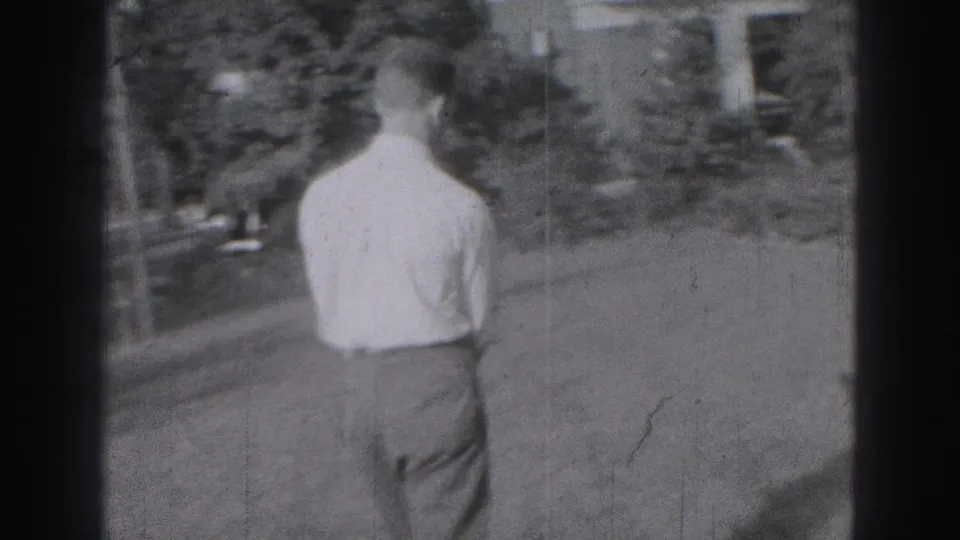 1975 Harrisburg frame showing a young man from behind walking across a grassy yard toward a residential building.