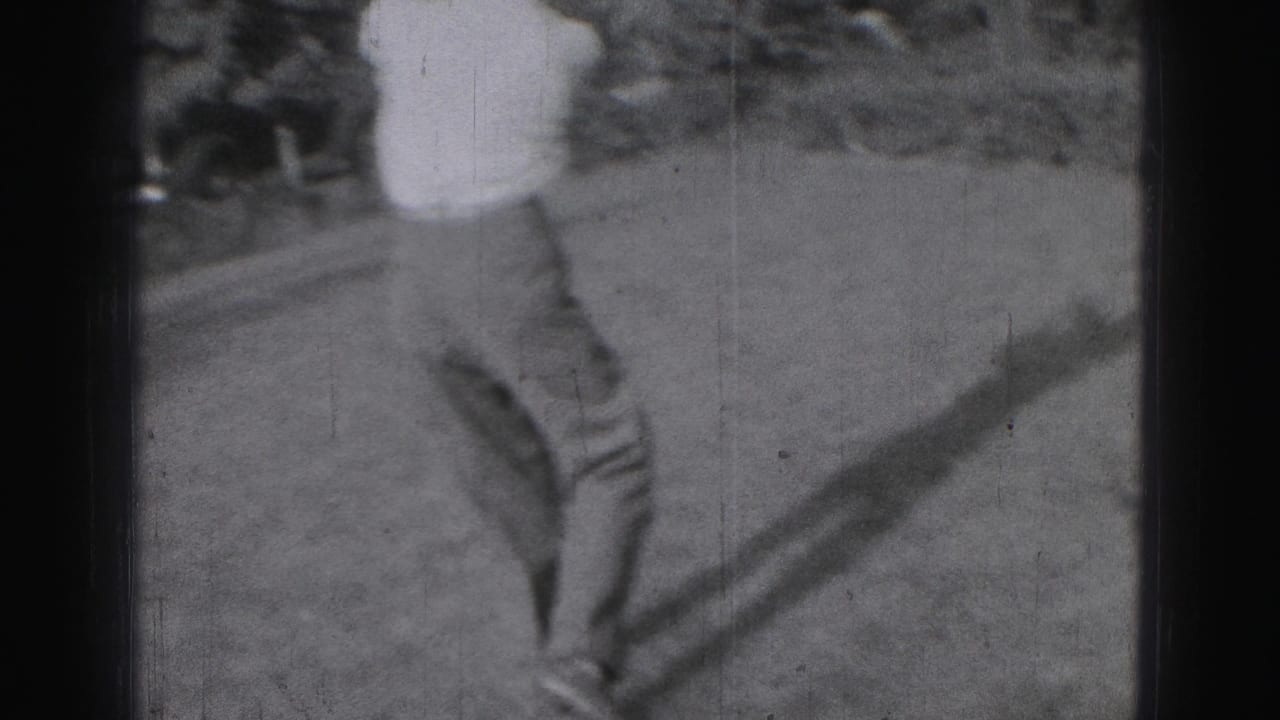 In 1975 Harrisburg home-movie footage, a young man practices a golf swing on a grassy area with trees behind him.