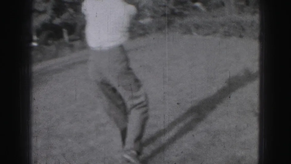 In 1975 Harrisburg home-movie footage, a young man practices a golf swing on a grassy area with trees behind him.