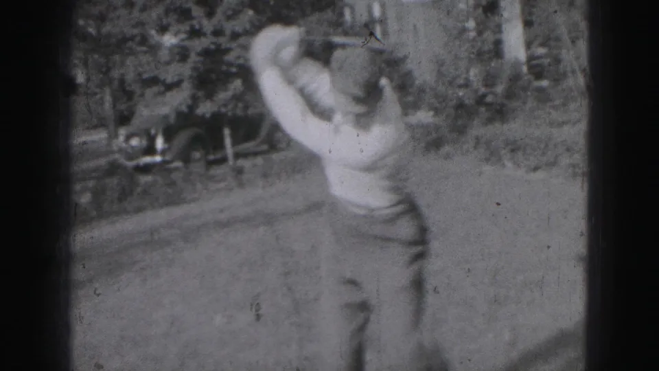 A young man completes a golf swing in 1975 Harrisburg footage, with a parked older car and trees in the background.