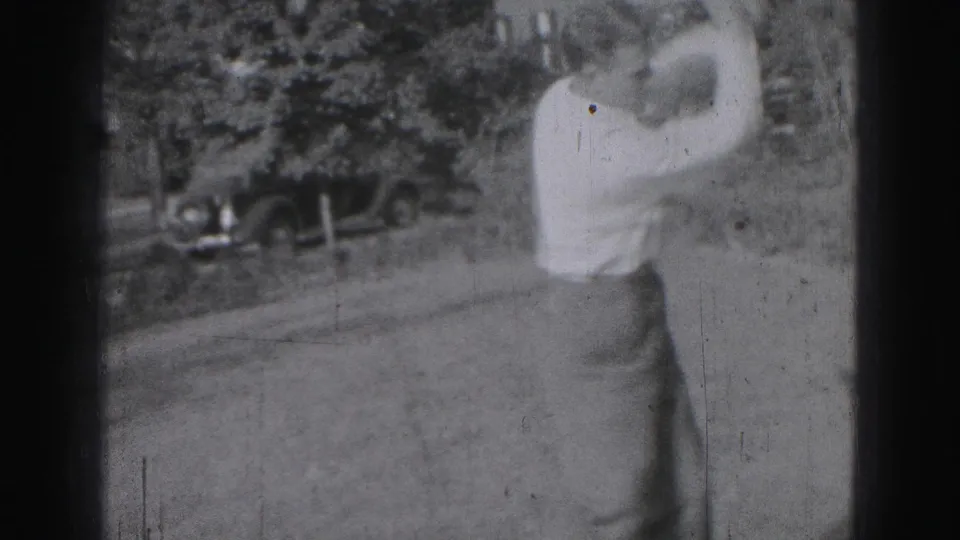 In Harrisburg 1975 home-movie footage, a young man in a light shirt prepares a golf swing outdoors near trees and a parked car.