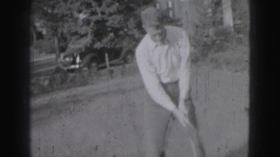 A young man in Harrisburg, Pennsylvania, bends into a golf stance with a club in black-and-white 8mm footage from 1975.