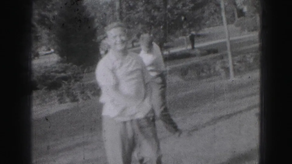 Two adult men in 1975 Harrisburg home-movie footage stand close together outdoors in a grassy setting.