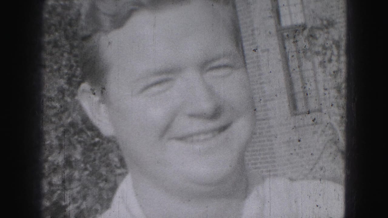 Close-up of a smiling adult man in 1975 Harrisburg home-movie footage, with shrubs and brickwork behind him.