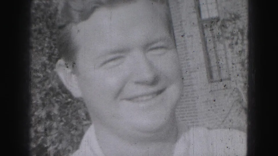 Close-up of a smiling adult man in 1975 Harrisburg home-movie footage, with shrubs and brickwork behind him.