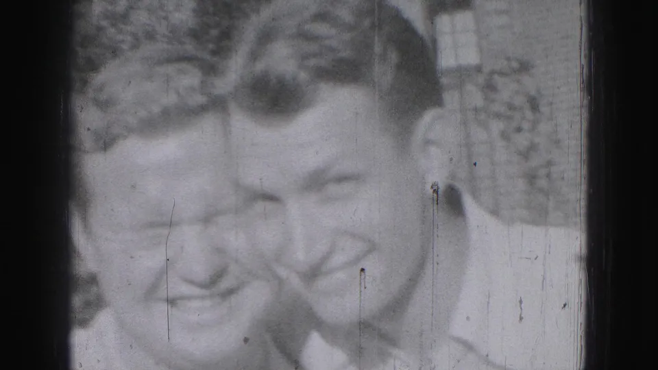 Two adult men smile close to the camera in 1975 Harrisburg home-movie footage, with a building and shrubs behind them.
