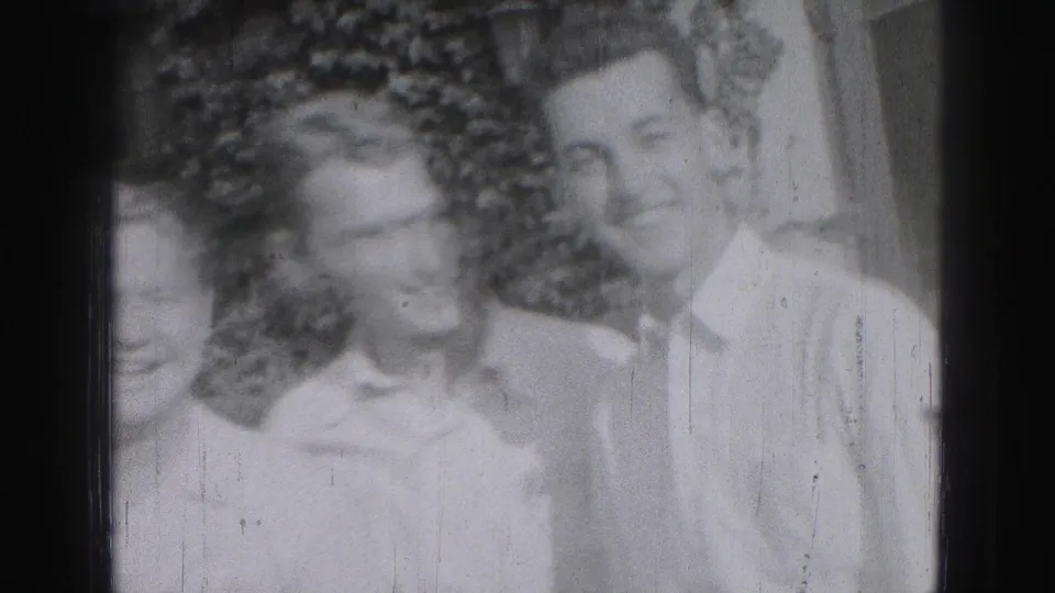 Two young men in light shirts smile in a 1975 Harrisburg outdoor home-movie frame, with a partial face at the edge.