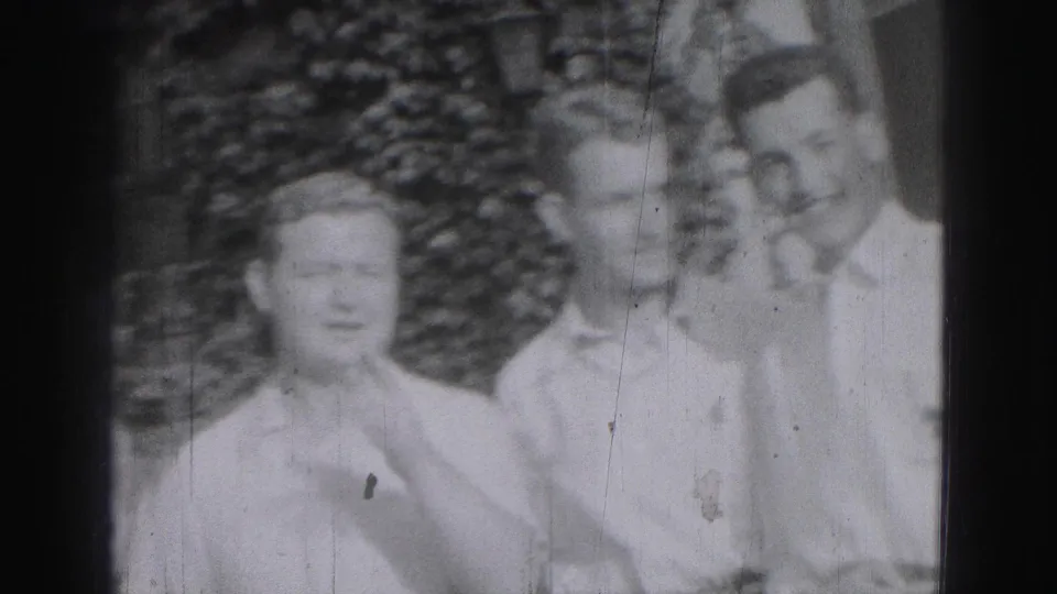 In a Harrisburg 1975 home-movie frame, three young men stand by foliage while one raises a hand to his face.