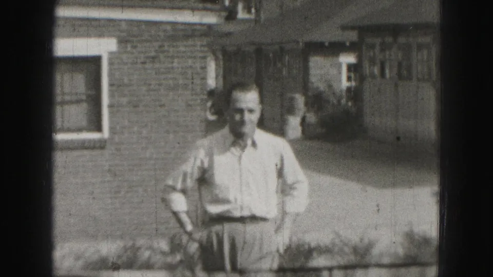 Adult man in a light shirt and trousers stands before a brick house in 1975 Harrisburg home-movie footage.