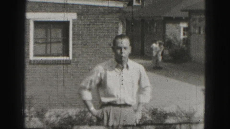 Older man in a light shirt stands outside a brick building in Harrisburg, Pennsylvania, in 1975 home-movie footage.