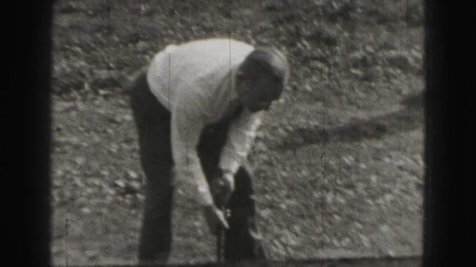 Man bends over on a gravel surface in Harrisburg, appearing to handle a stick or club in 1975 home-movie footage.