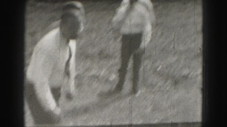 Two adult men appear outdoors in Harrisburg, with one bending near the ground in soft 1975 8mm footage.