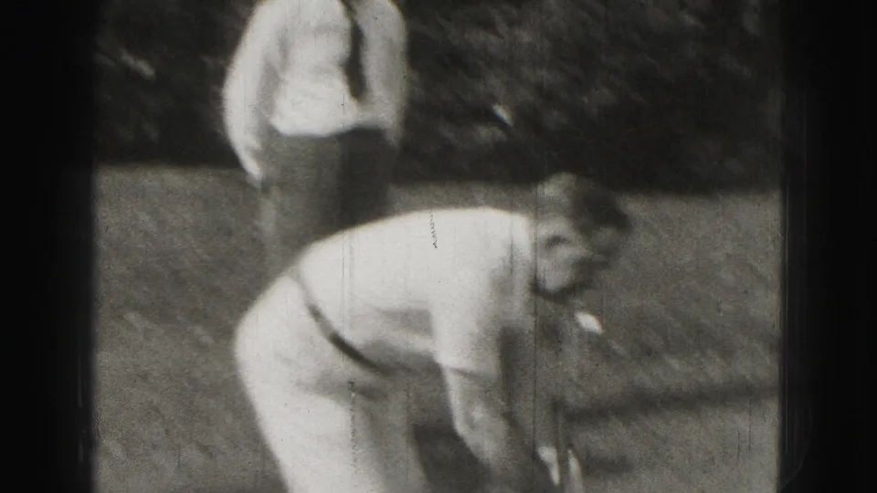 In the supplied 1975 Harrisburg home-movie context, a man bends over a croquet shot in a backyard while another adult stands nearby.