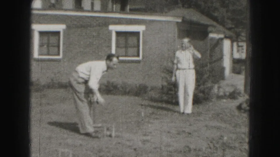 A backyard croquet player bends toward the ground while a second man watches near a brick house with white-trimmed windows.