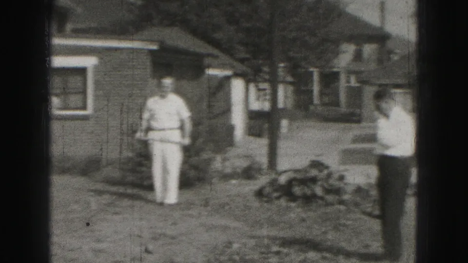A man stands in a residential yard near a brick house, with another adult partly visible along the edge of the home-movie frame.