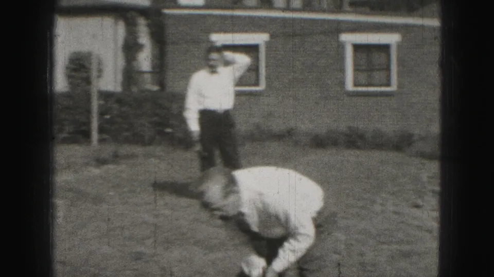1975 Harrisburg home-movie frame showing men in a suburban backyard, one bending to play croquet while another watches near a house.