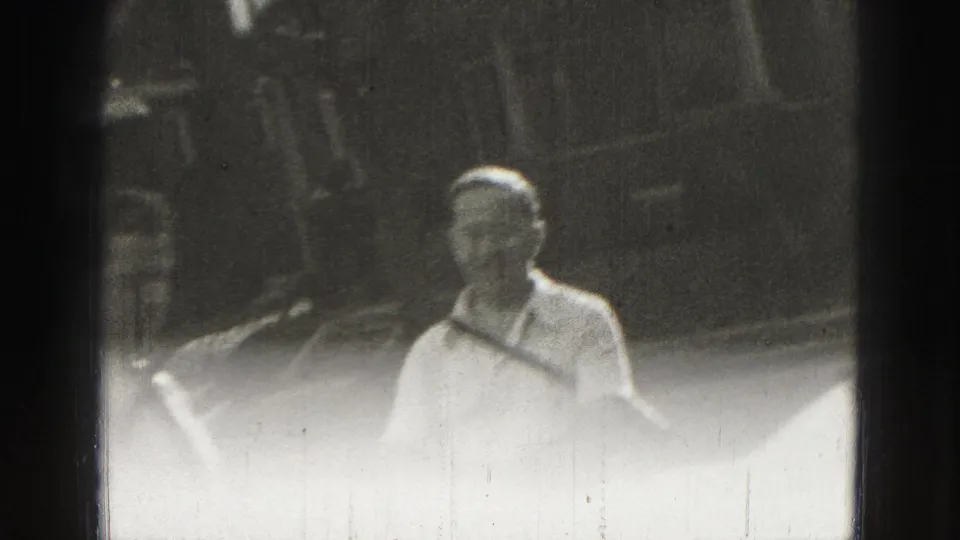 Adult man in light summer clothing stands in the Harrisburg home-movie scene, framed softly against an outdoor yard background.