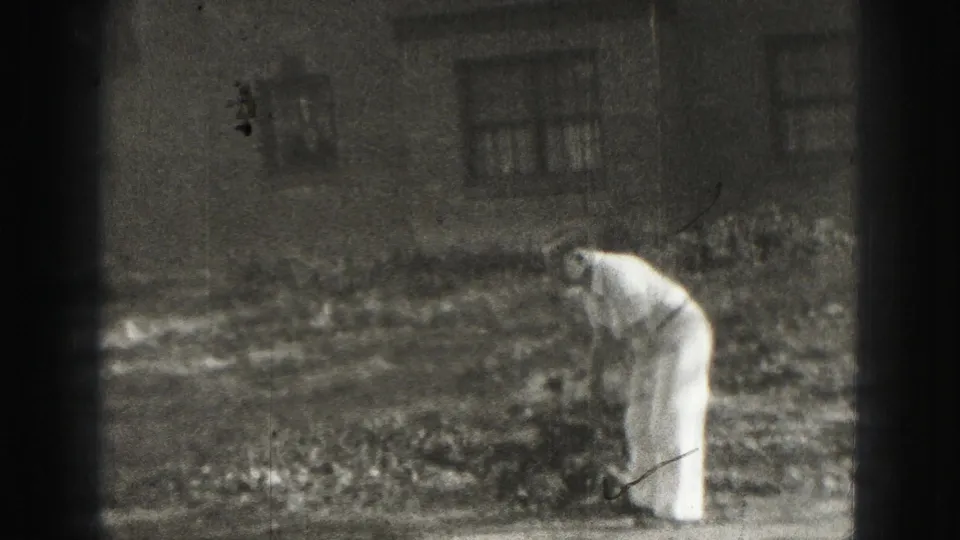 An adult bends over with a club or mallet in a Harrisburg yard, suggesting a casual lawn game in the 1975 home movie.