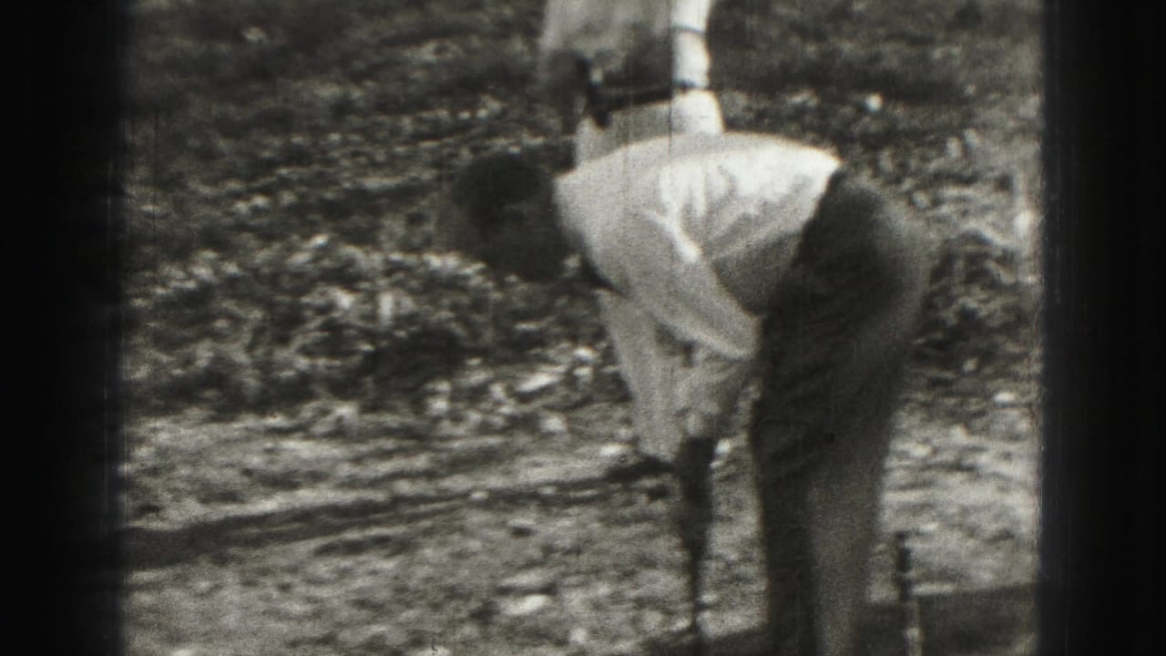 Golfer bends over a club while another person stands behind on a grassy course.