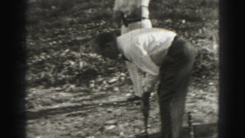 1975 Harrisburg home-movie frame showing a golfer in a light shirt bending over a club as another person stands nearby.