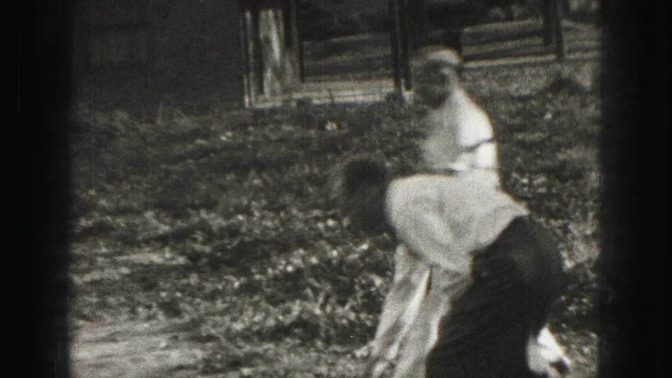 Home-movie frame from Harrisburg in 1975 showing a golfer near a standing observer with a structure in the background.