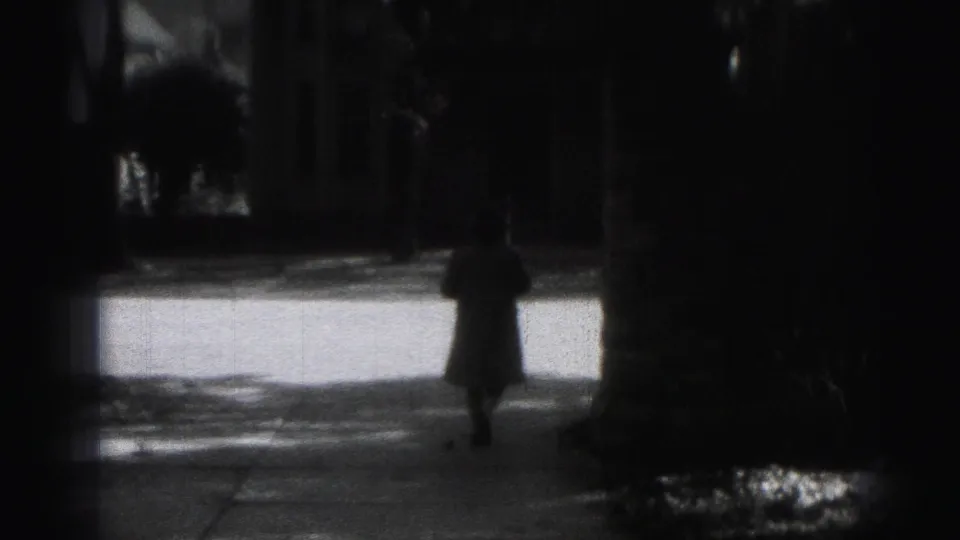 1975 Harrisburg home-movie frame with a small silhouetted pedestrian beneath dark trees near a bright sidewalk.