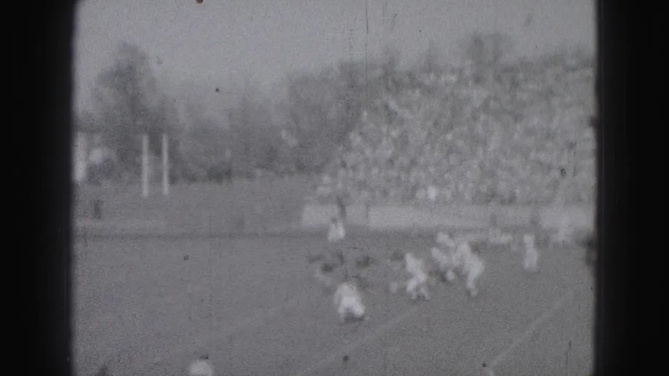 Distant 1975 home-movie view of a football play in Harrisburg, Pennsylvania, with players on the field and a large crowd in the stands.