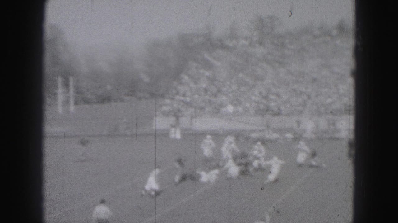 Distant 8mm frame of football players converging during play, with a packed stadium crowd visible behind the field.
