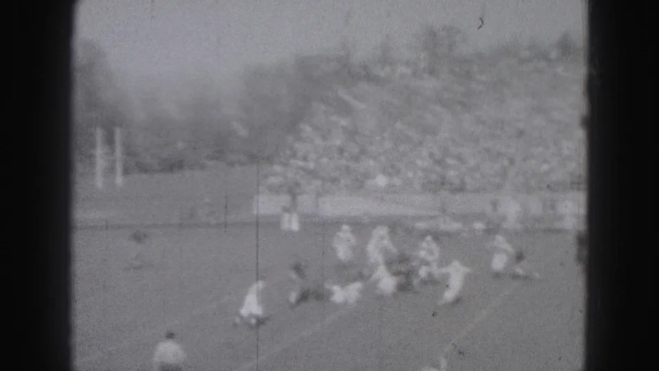 Distant 8mm frame of football players converging during play, with a packed stadium crowd visible behind the field.