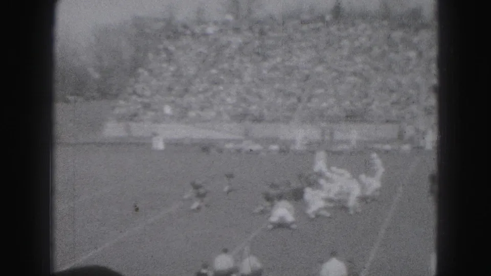 Grainy home-movie view of a football tackle or pileup on the field, with Harrisburg stadium spectators in the background.